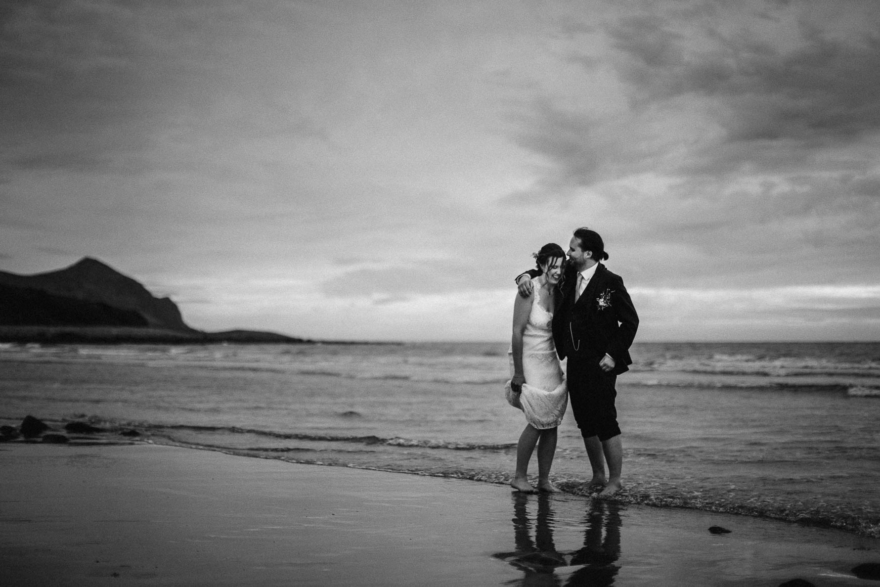 Black and white shot of the groom with his arm around the bride's shoulder as they are stood barefoot on the beach as the water is coming up over their feet