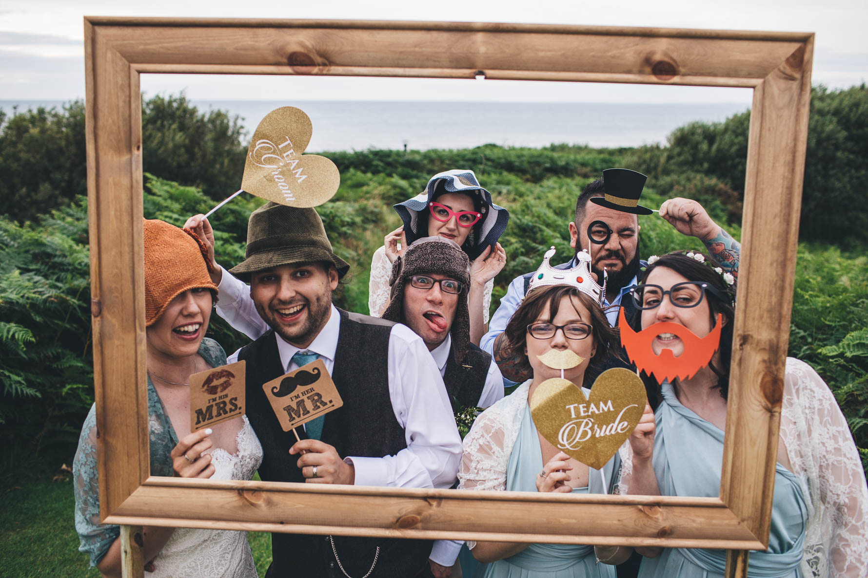 Wedding guests with various glasses and props stood behind a large picture frame pulling faces and smiling
