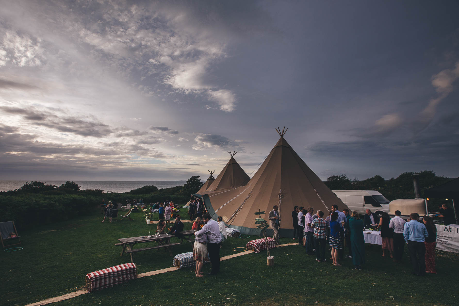 Shot of three large tipi's on a lawn with wedding guests in and around the tipi's as the sky is starting to cloud over in the early evening