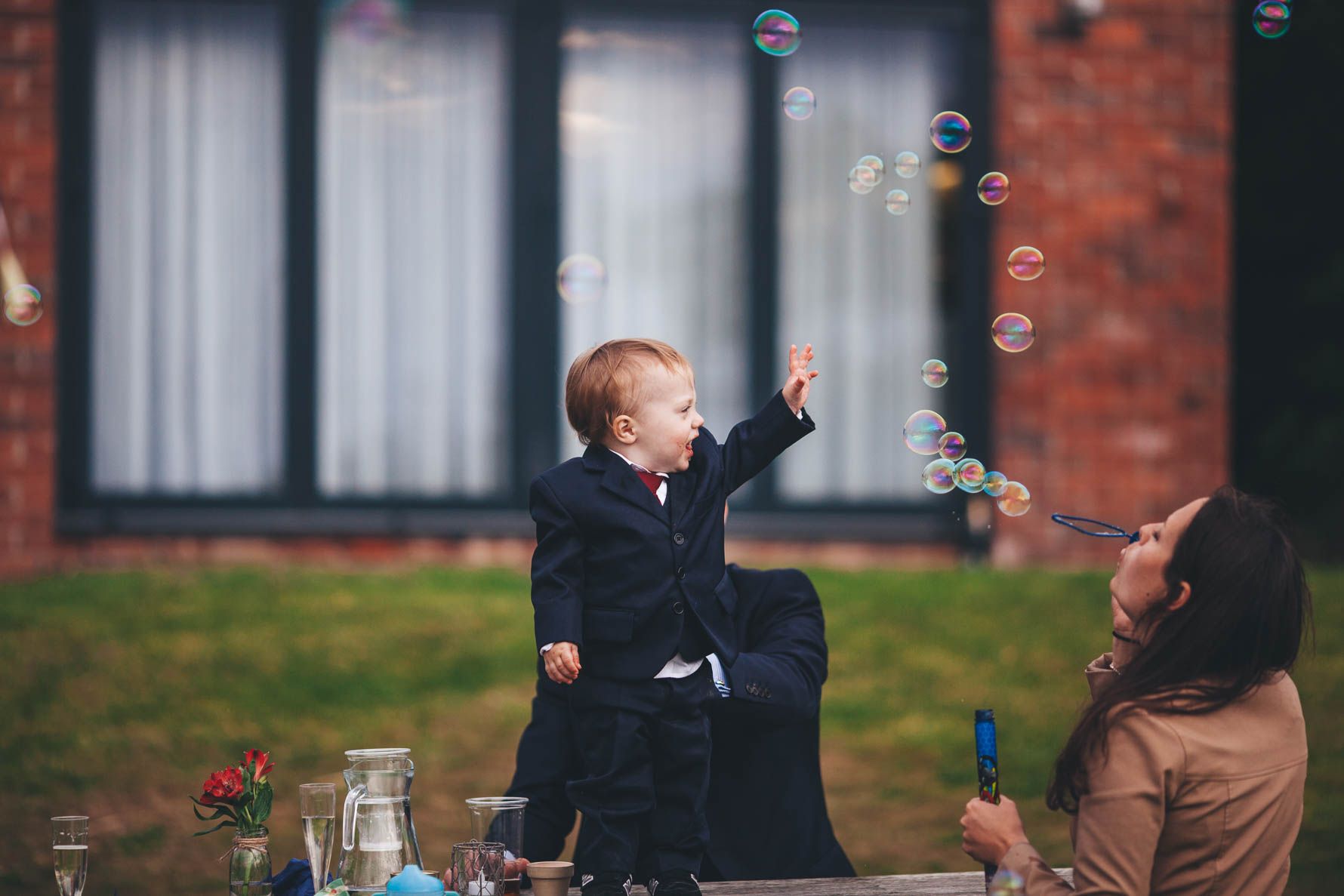 Young boy stood on a table trying to catch bubbles which a woman is blowing in front of him