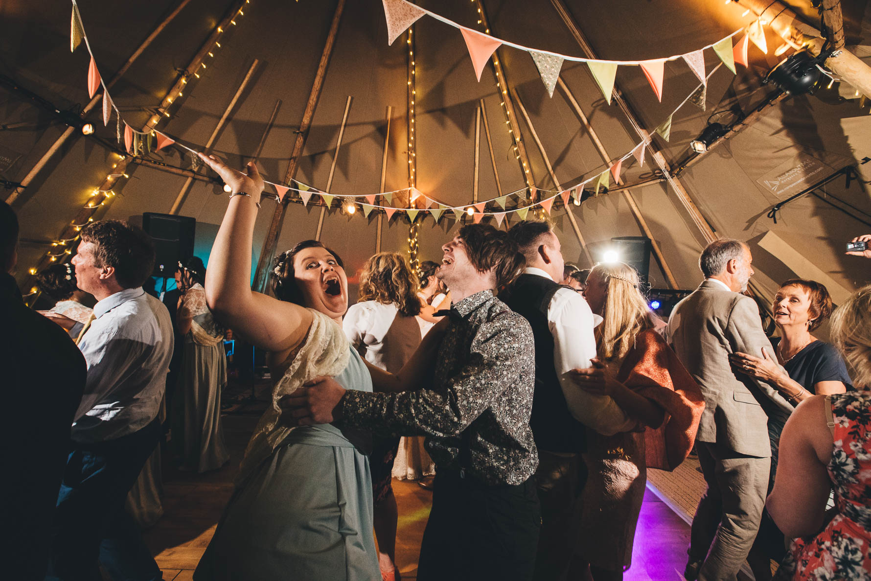 Wedding guests dancing inside a tipi with a bridesmaid with her har in the air and mouth wide open as her partner has his arms around her waist and is laughing