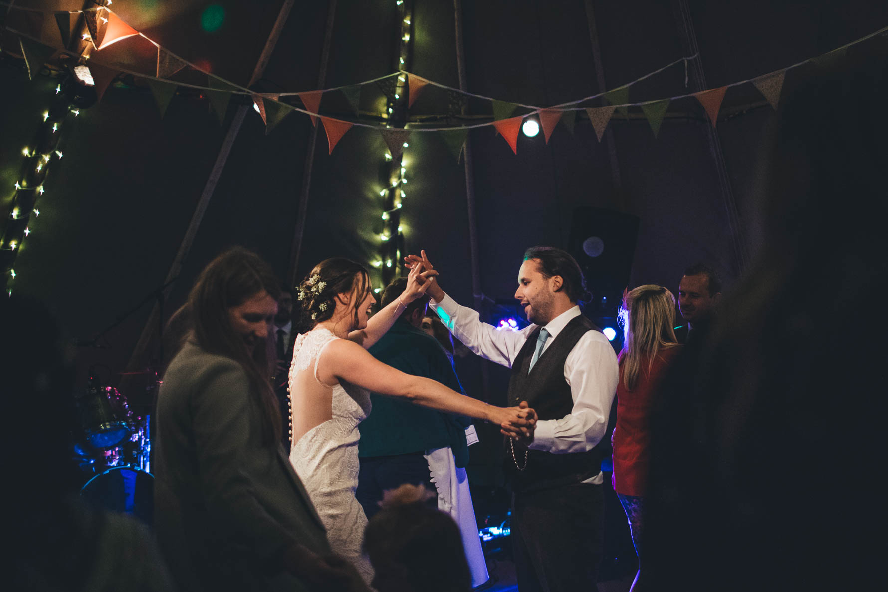 Bride and groom with their hands togetherin the air as they dance inside a tipi with bunting and fairylights on the supports of the tipi