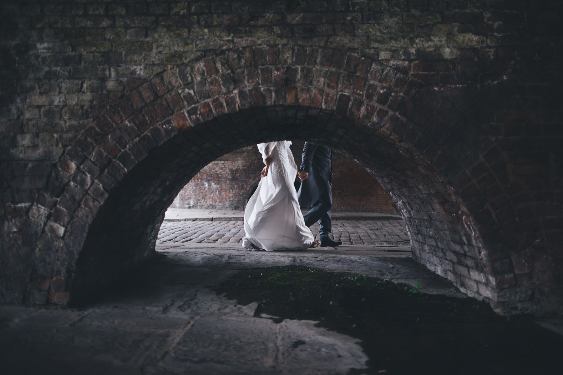 couple walk behind brick arch portrait