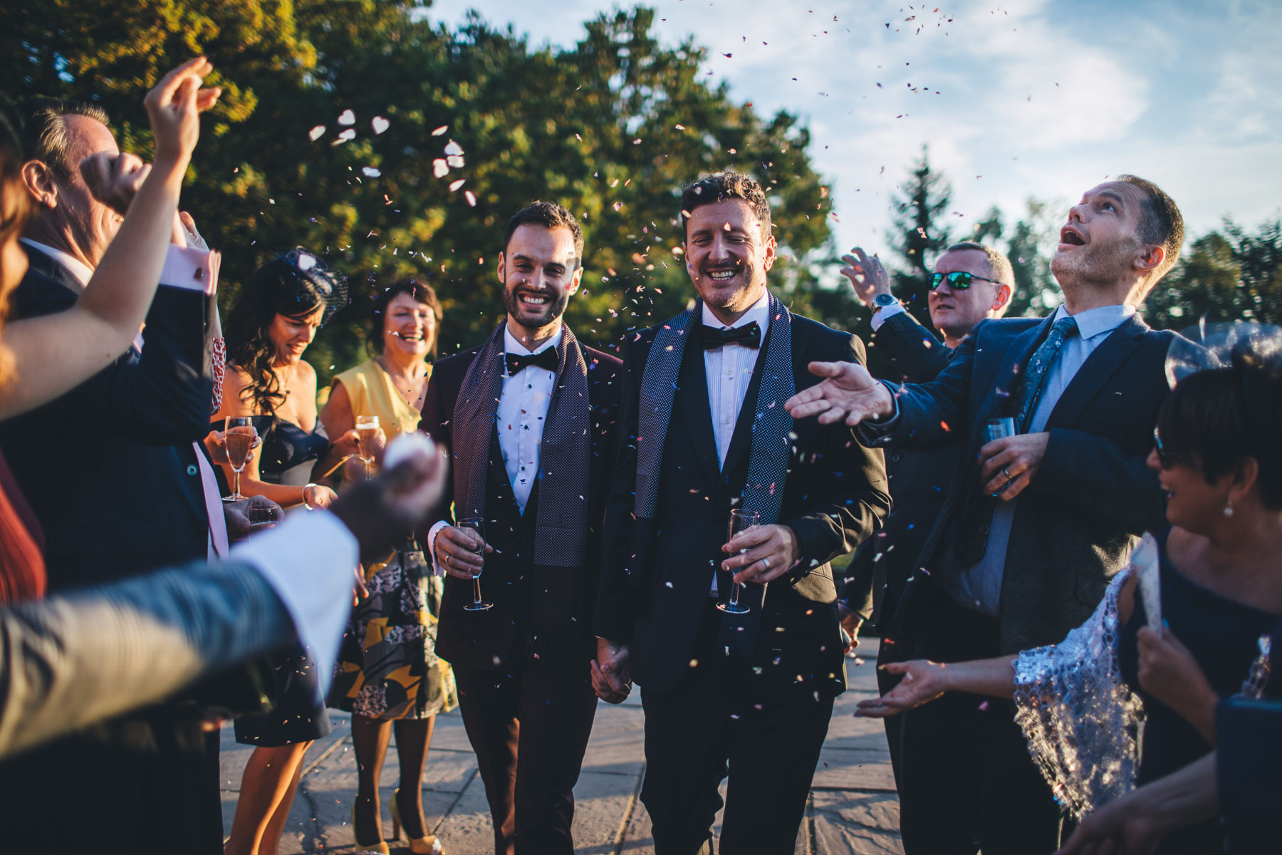 two groom wearing bow ties walk through a procession of confetti