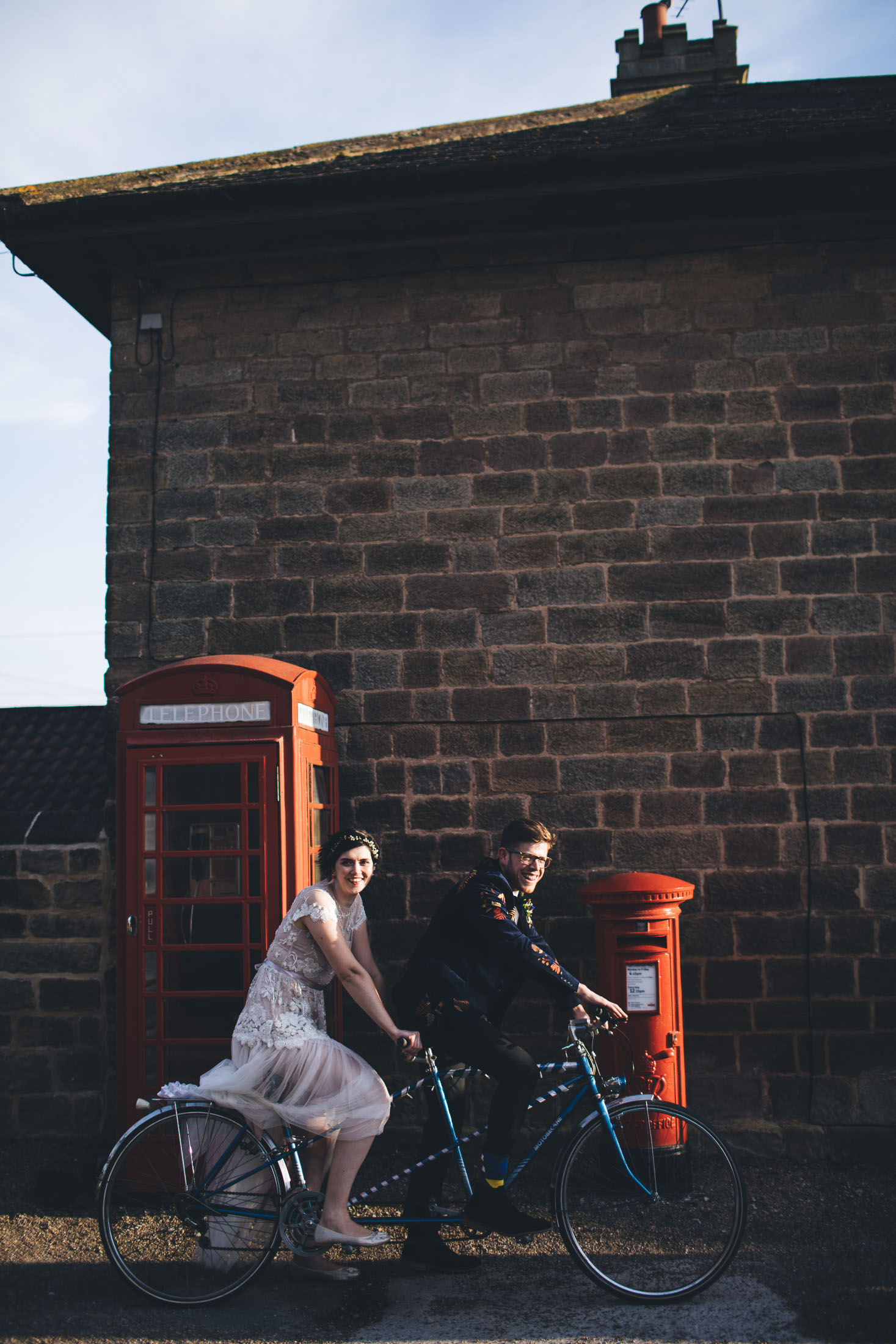 couple pose next to red post box and telephone box on their tandom bike