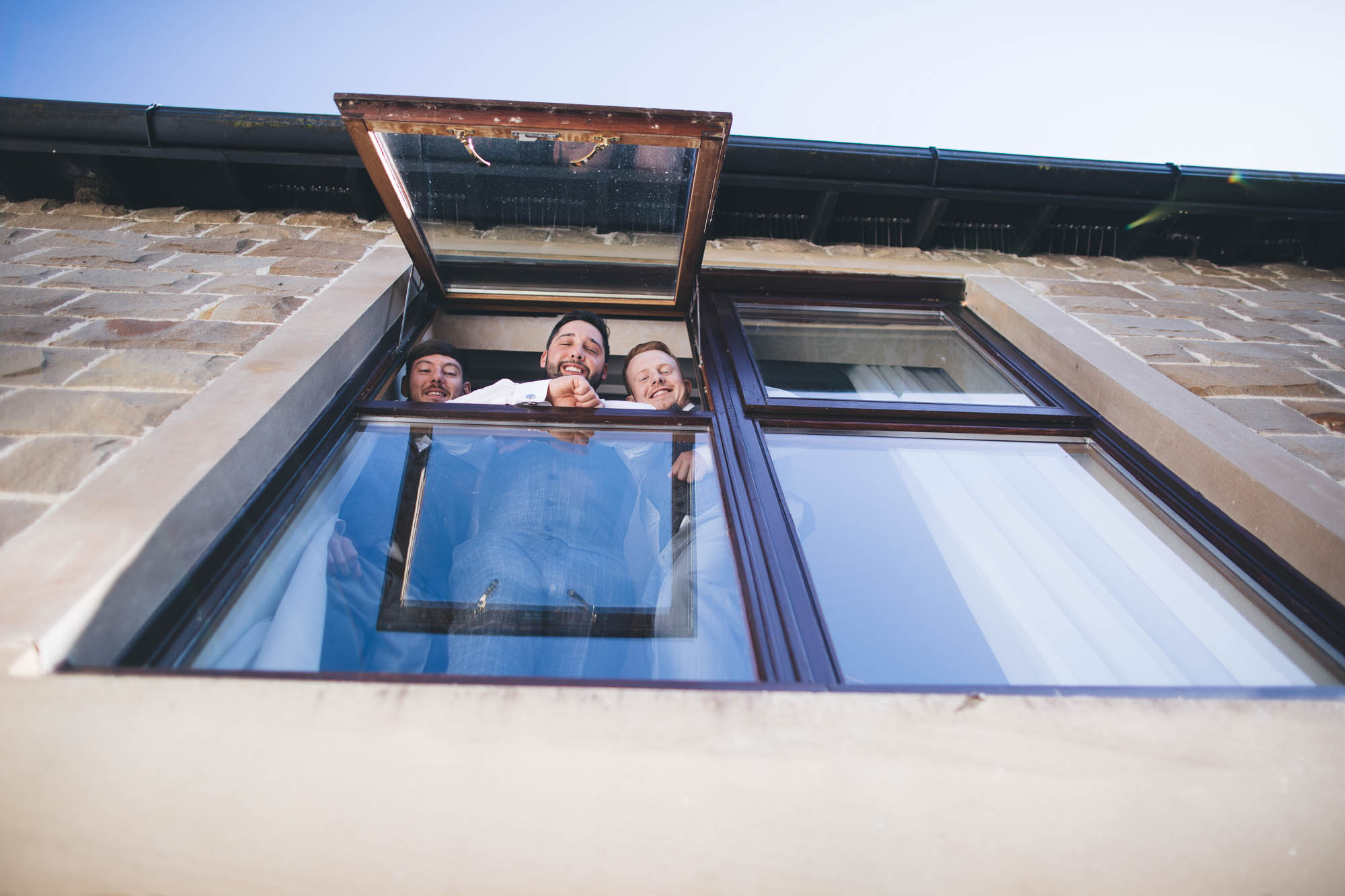 Groom and Groomsmen look down from window above cheekily on morning of wedding