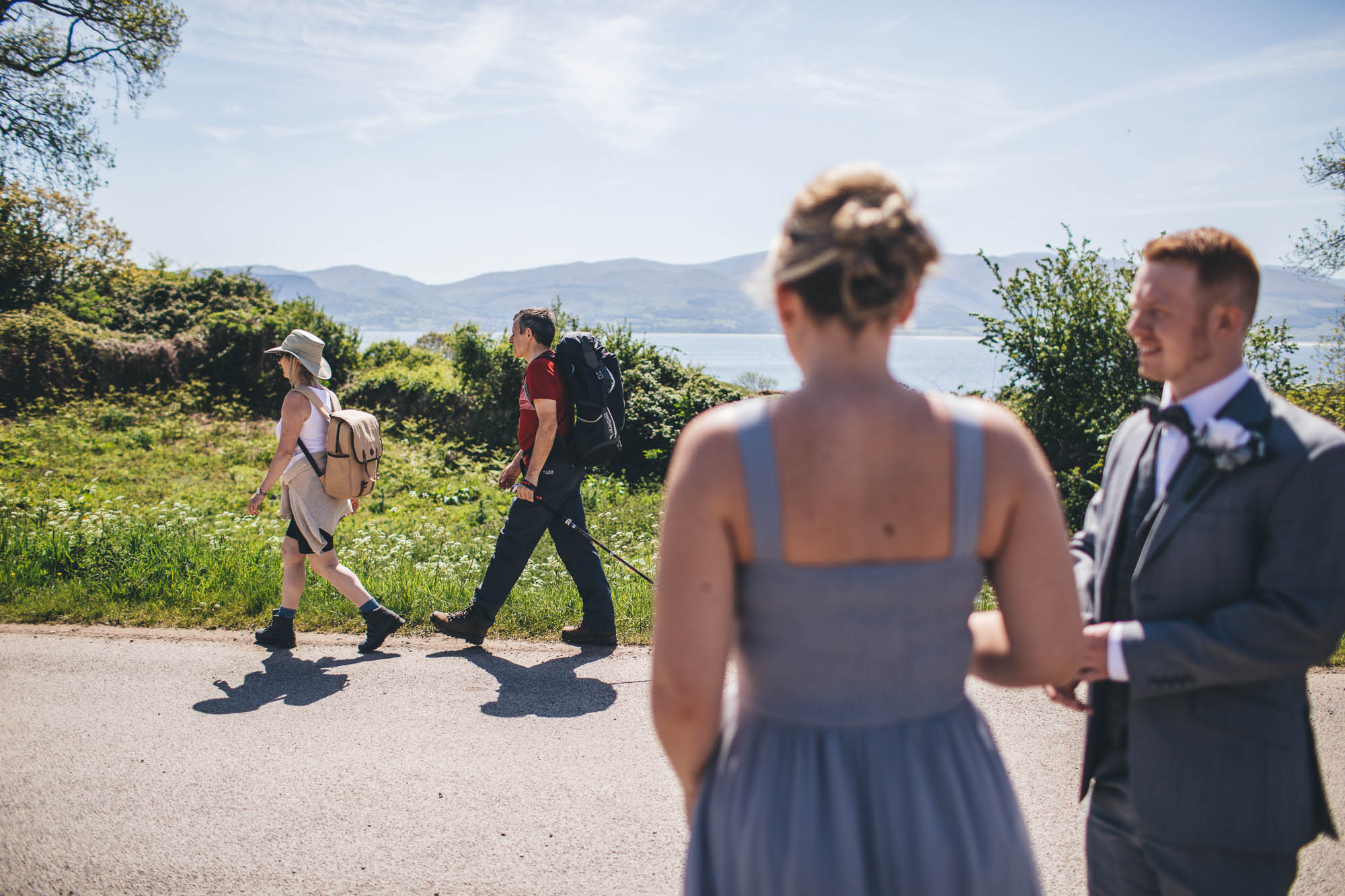 Wedding Guests watch members of public walk past on a hike with Anglesey coastline in background as they wait for Bride and Groom to arrive