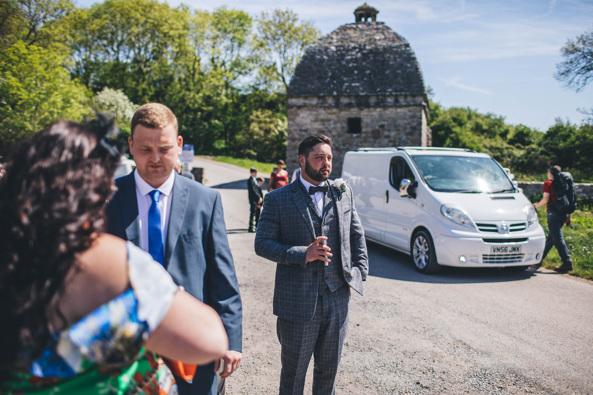 Groom stands in road amongst wedding guests in anticipation of his bride arriving at the Church