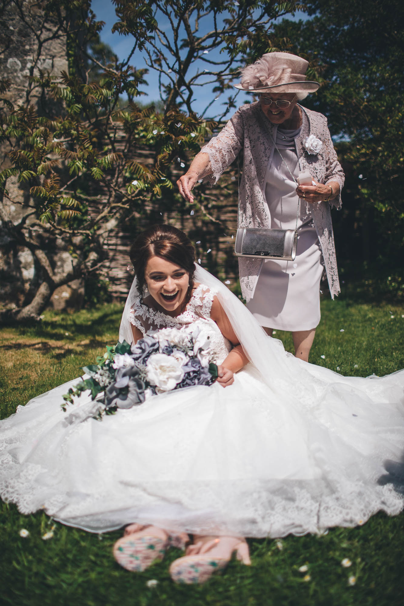 Bride sits on green grass whilst her Grandmother gleefully throws confetti over the Bride's head