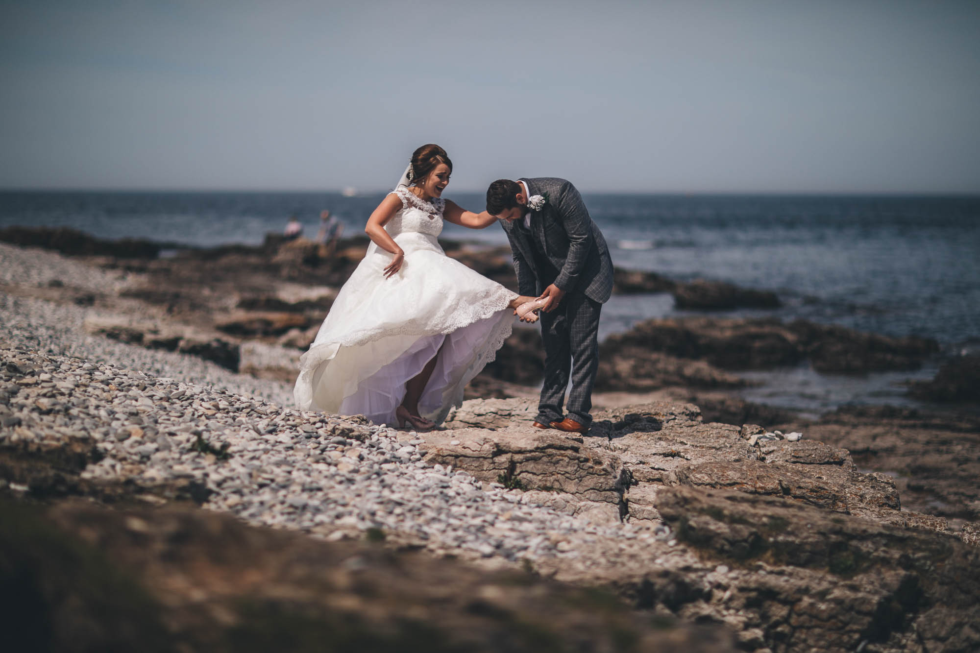Bride holds on to Groom to balance as they walk across rocky part of Anglesey coastline
