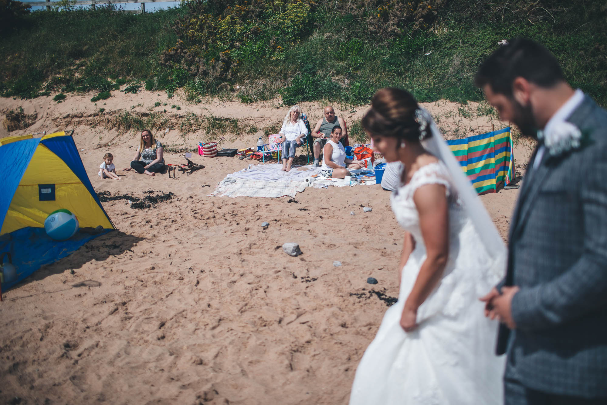 Just married couple are out of focus in the foreground of the shot on the beach to draw focus to the family looking onward in the background with their tent and windbreaker