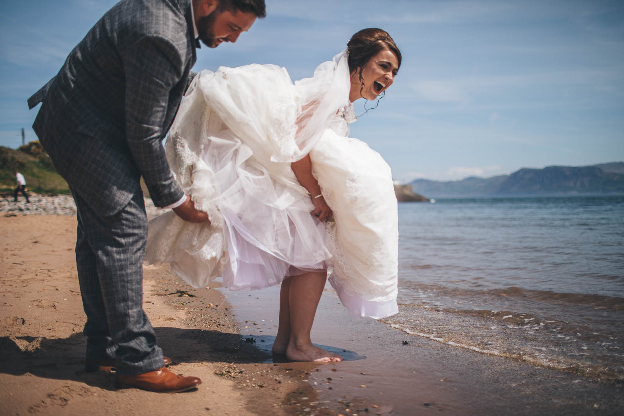 Groom helps to lift back of Bride's wedding dress as she laughs and goes for a paddle in the seaside