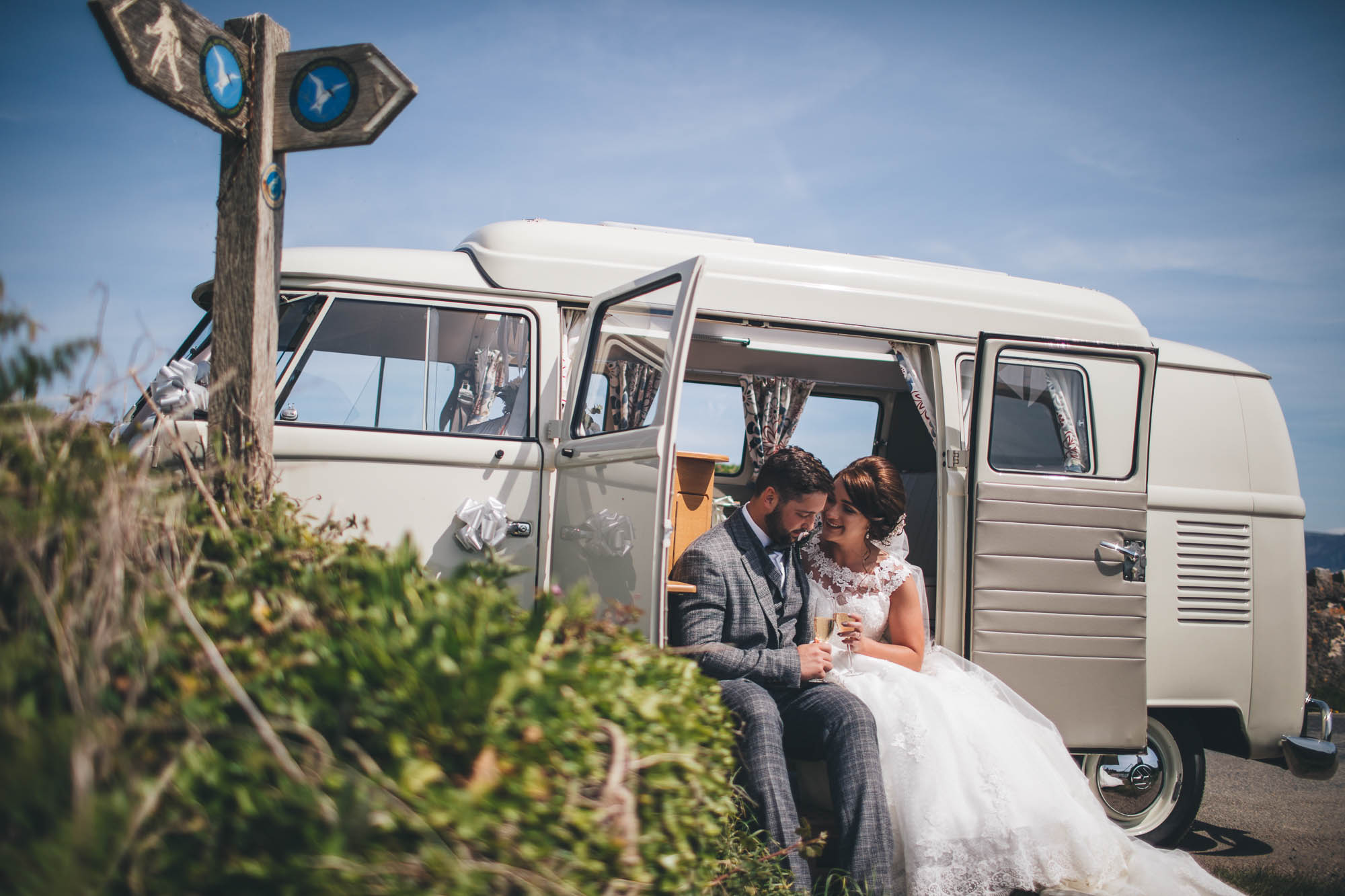 Newlyweds enjoy a cuddle and a glass of bubbly sat on the edge of vintage cream van by the Anglesey seaside