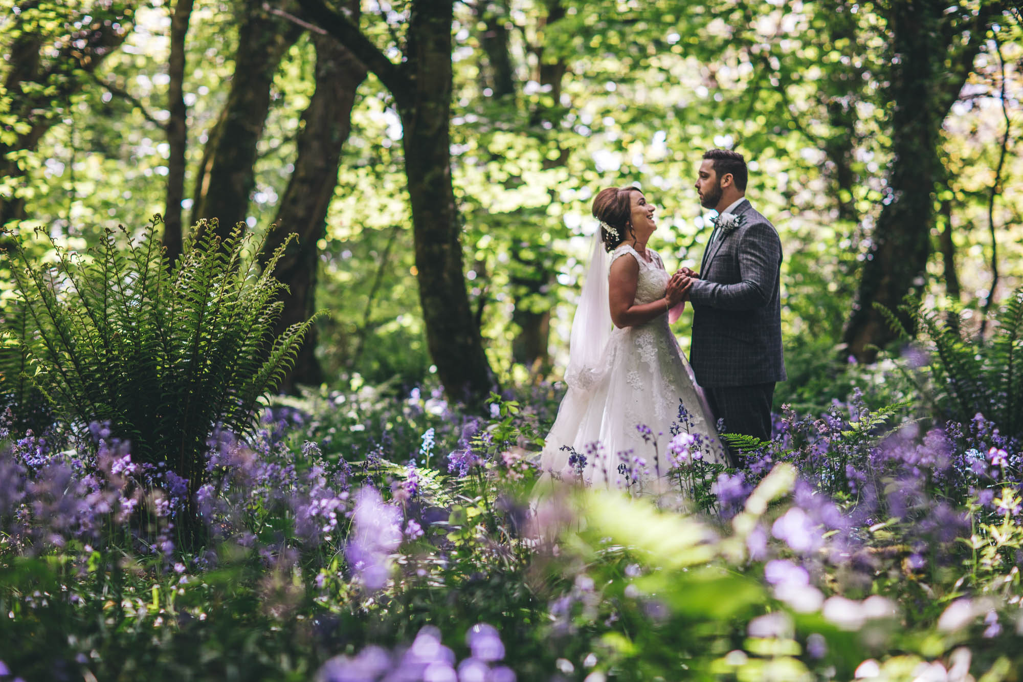 Groom making Bride laugh whilst they hold hands surrounded by greenery and purple flowers