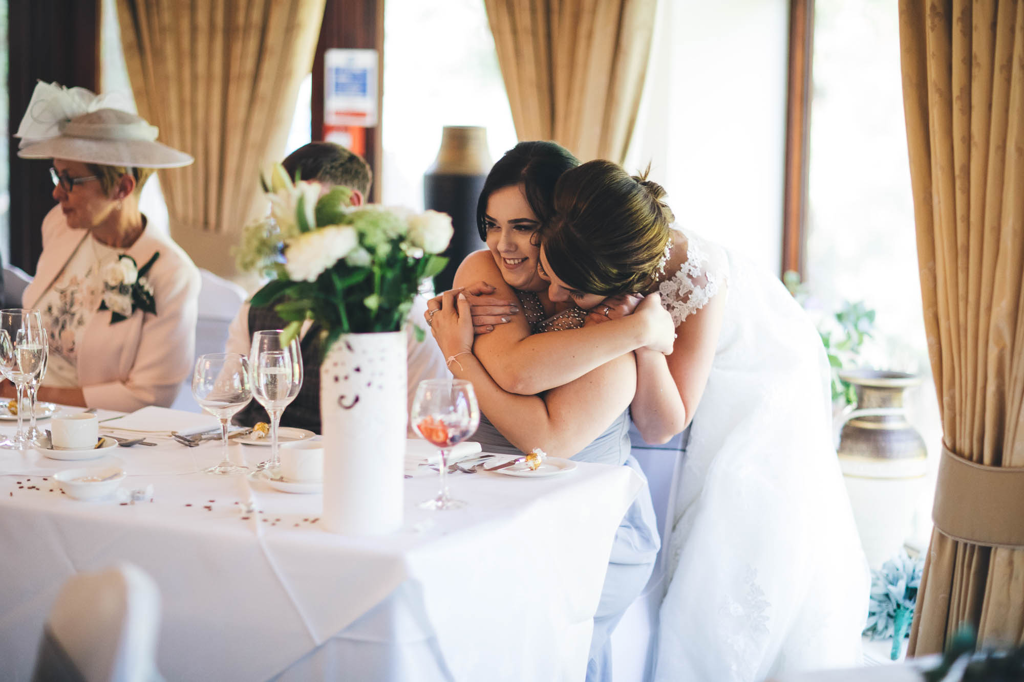 Bride embraces her maid of honour from behind at the top table