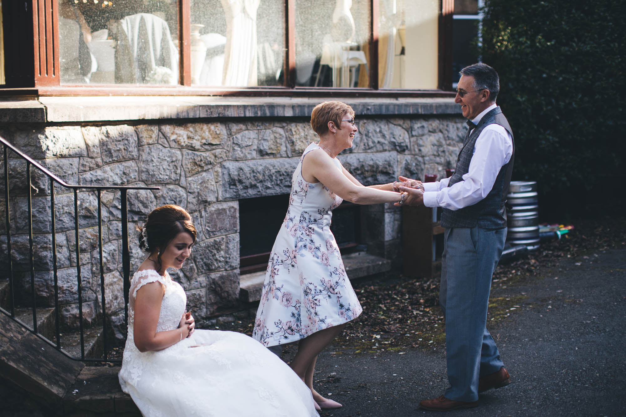 Bride sits on step while her parents dance with each other happily at wedding reception