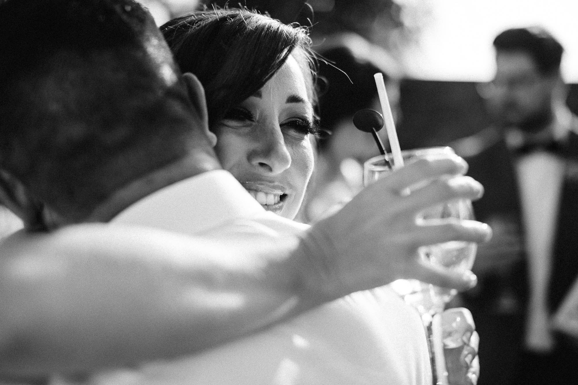 Black and shot of Bride receiving an embrace whilst she holds a drink