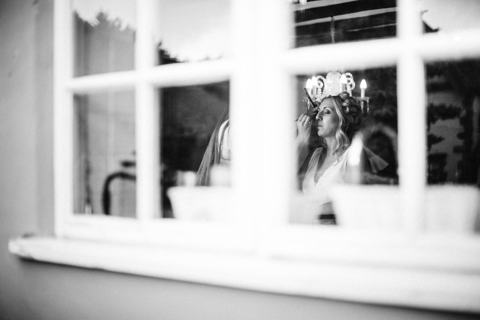 Black and white shot looking through window at Bride having her make up done