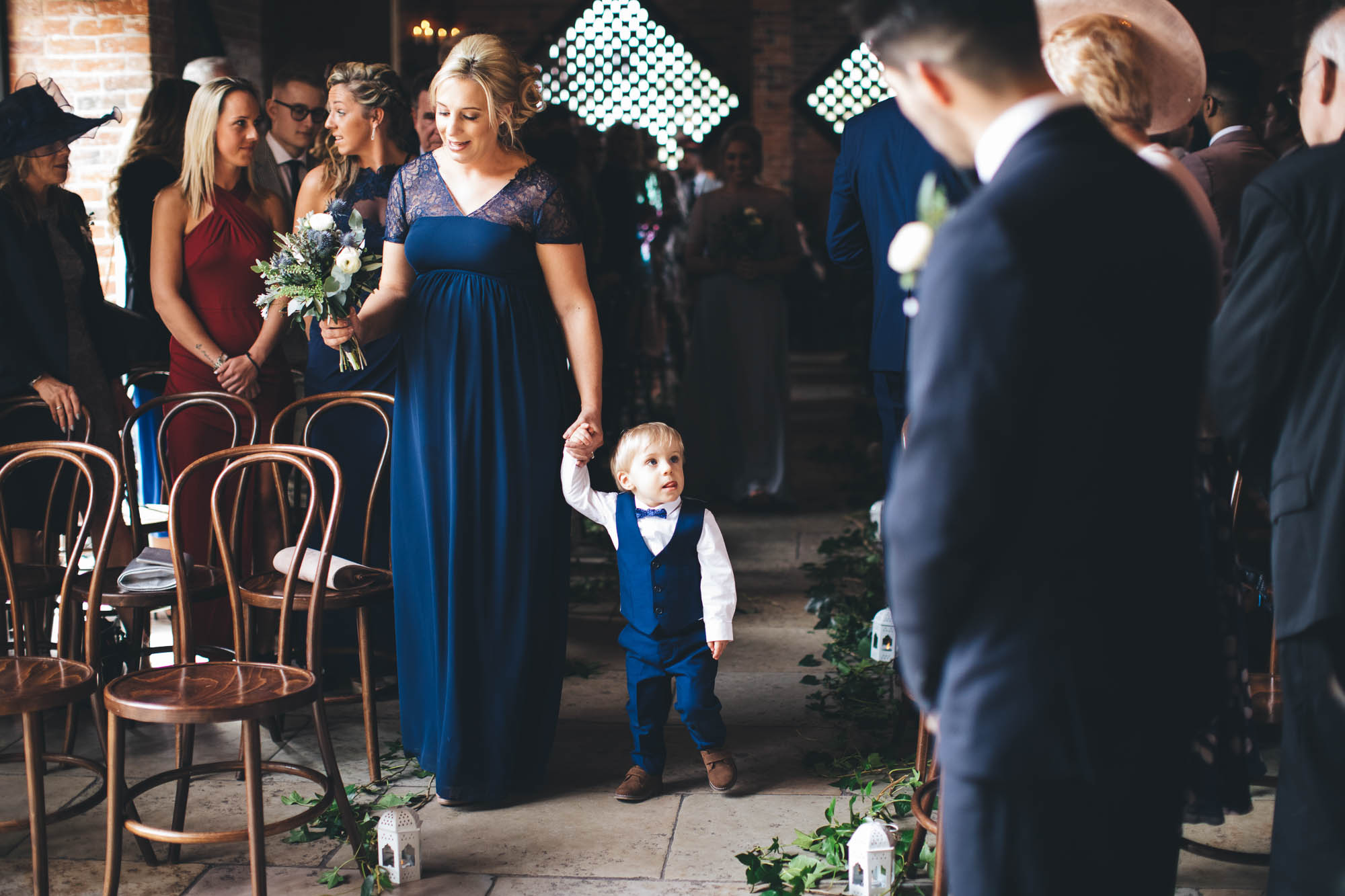 Maid of honour guides young boy down the aisle as guests wait arrival of Bride