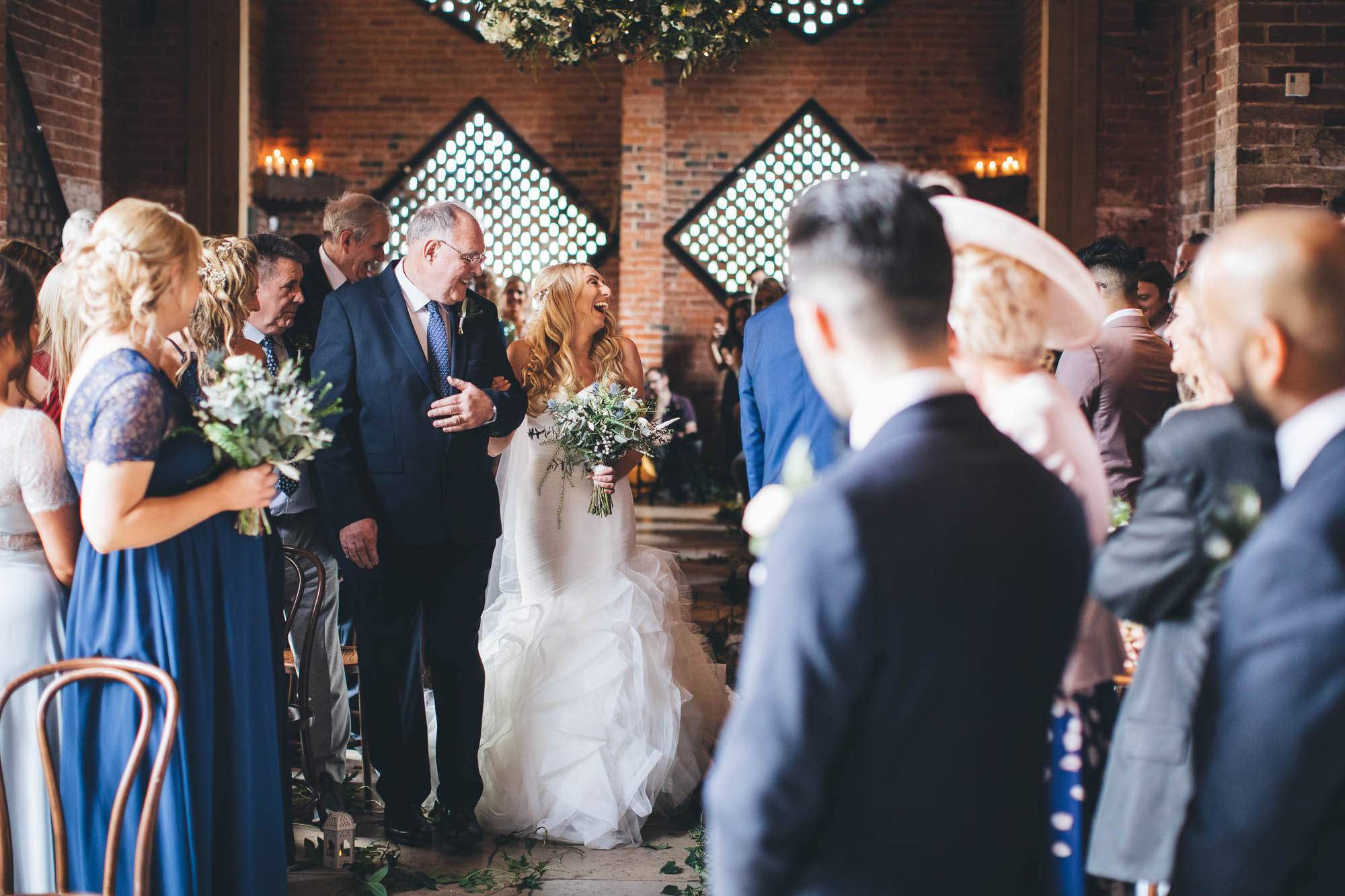 Father and Bride laugh with guests as they walk down the aisle in rustic barn
