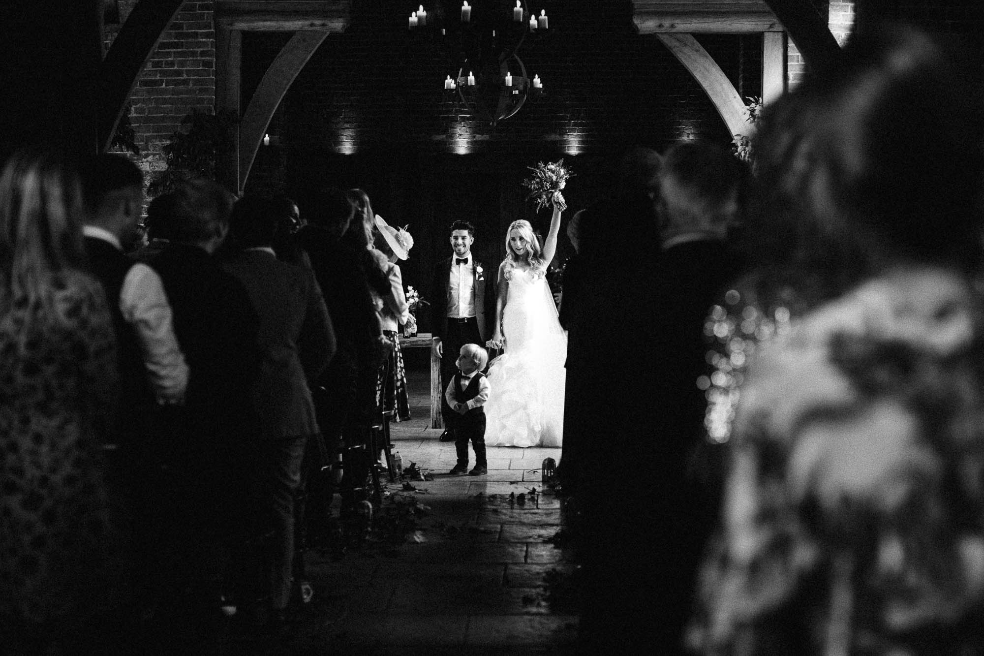 Black and white shot raises wedding bouquet as she walks out of the ceremony with her new husband