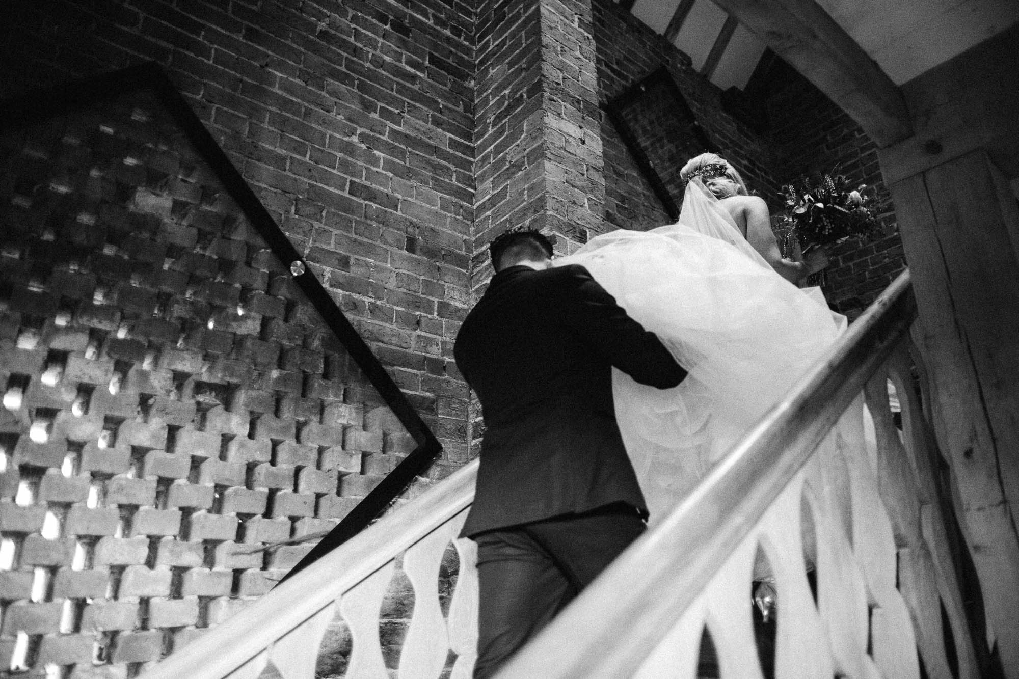 Black and white shot of groom helping to carry Brides wedding dress train up the stairs