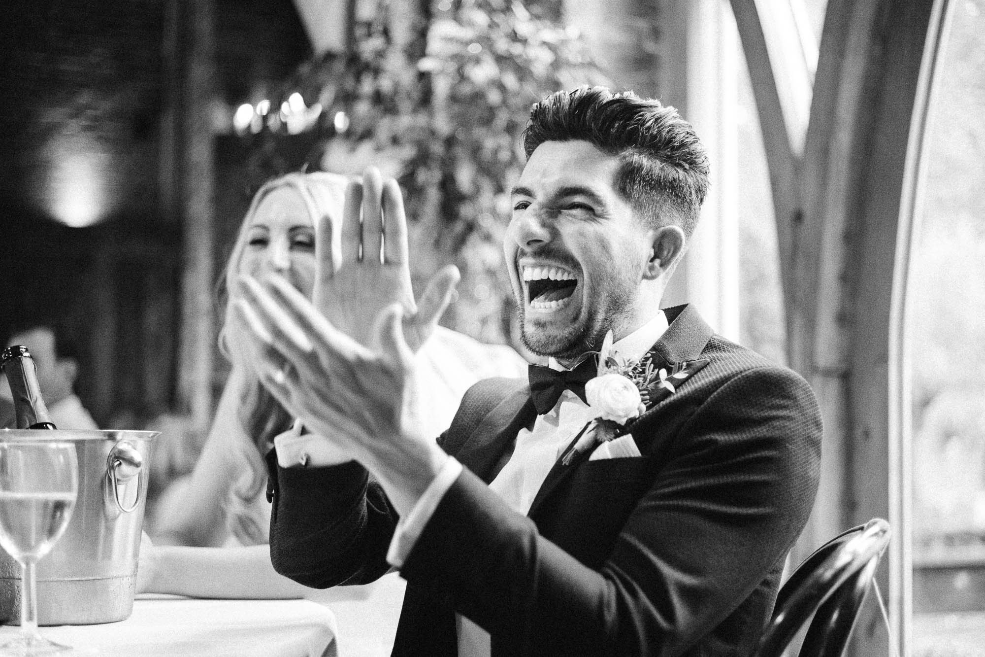 Monochrome shot of Groom laughing and clapping at wedding reception banter