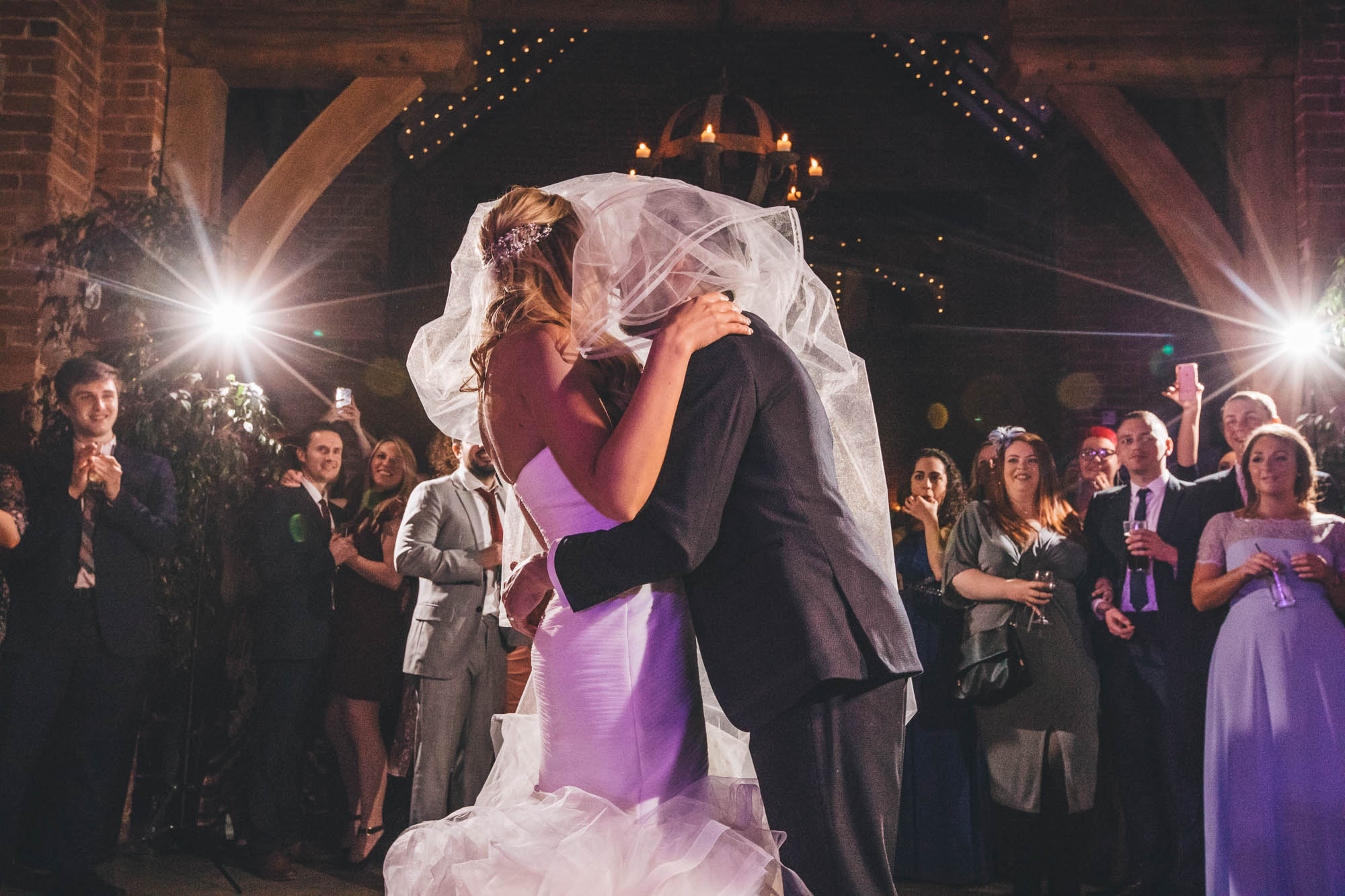Newlyweds share a kiss under the veil in rustic barn and guests look on
