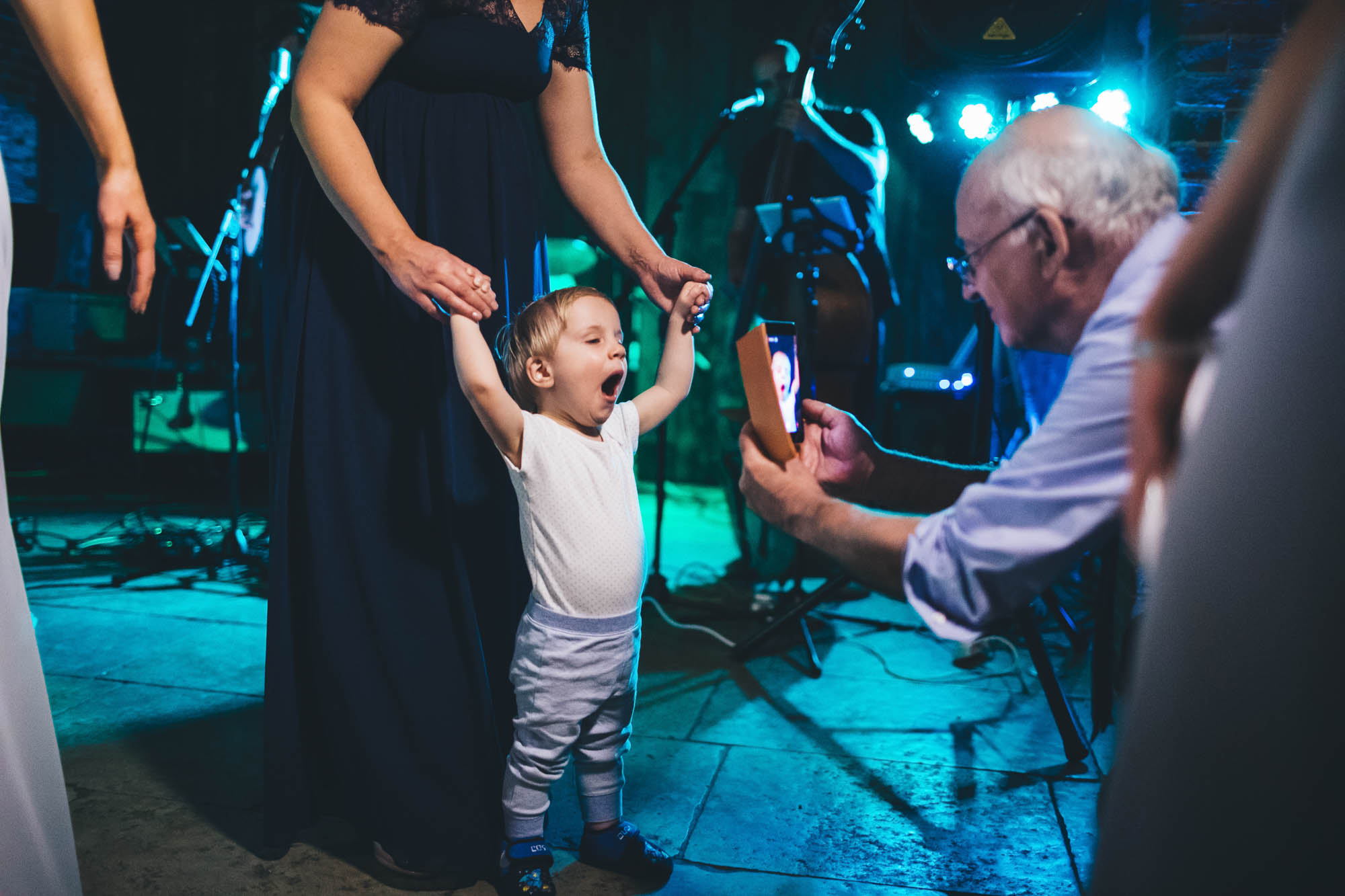 Young wedding guest yawns on the dancefloor as old man takes opportunity to get a photo of the cute moment