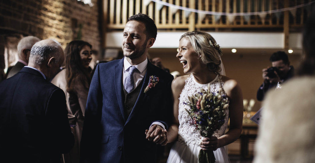 Bride and groom smile and hold hands as they leave the wedding barn as newlyweds