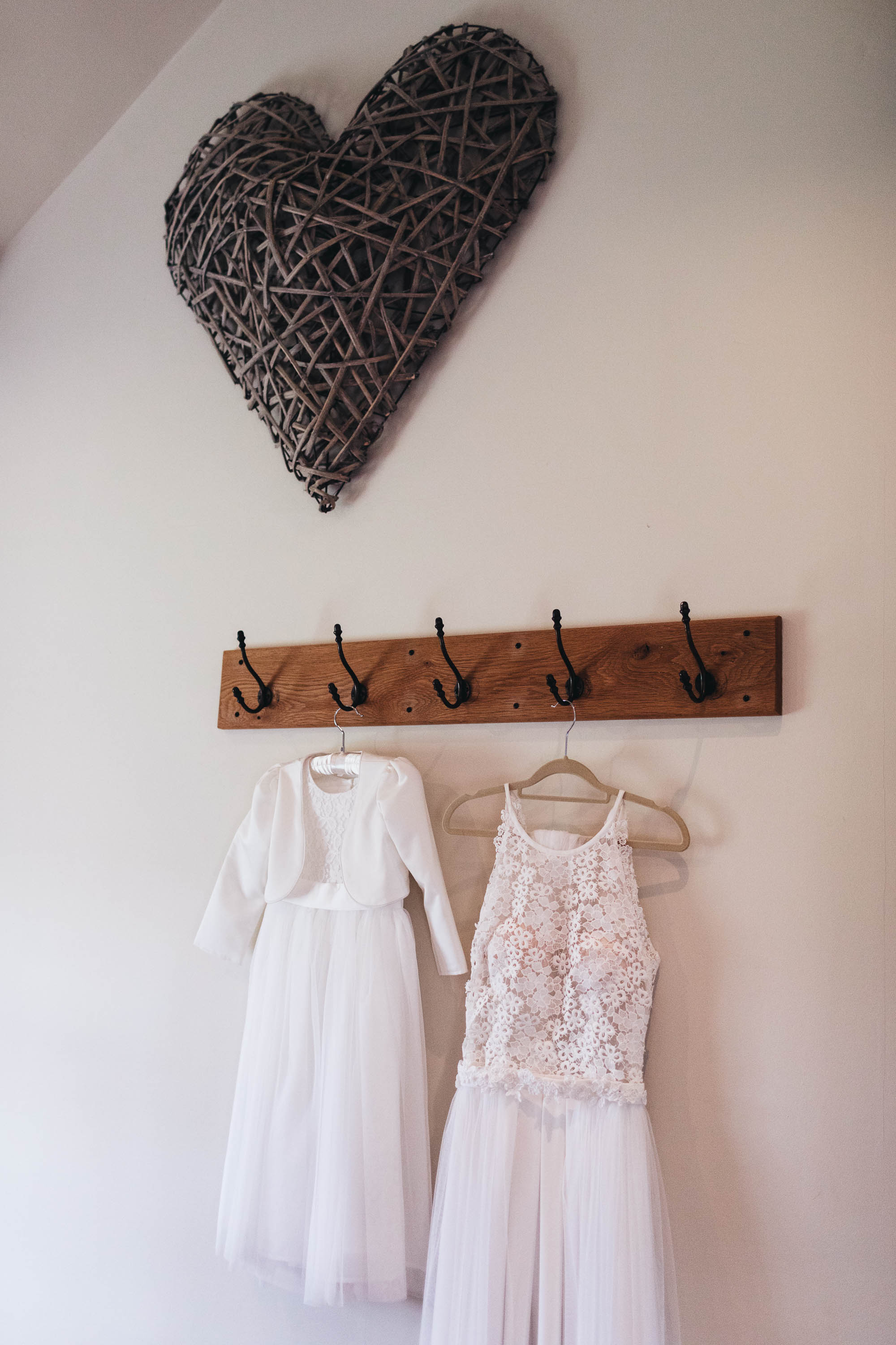 Wedding dress and flower girl dress hung up next to each other with a wooden thatched heart sculpture on the wall above