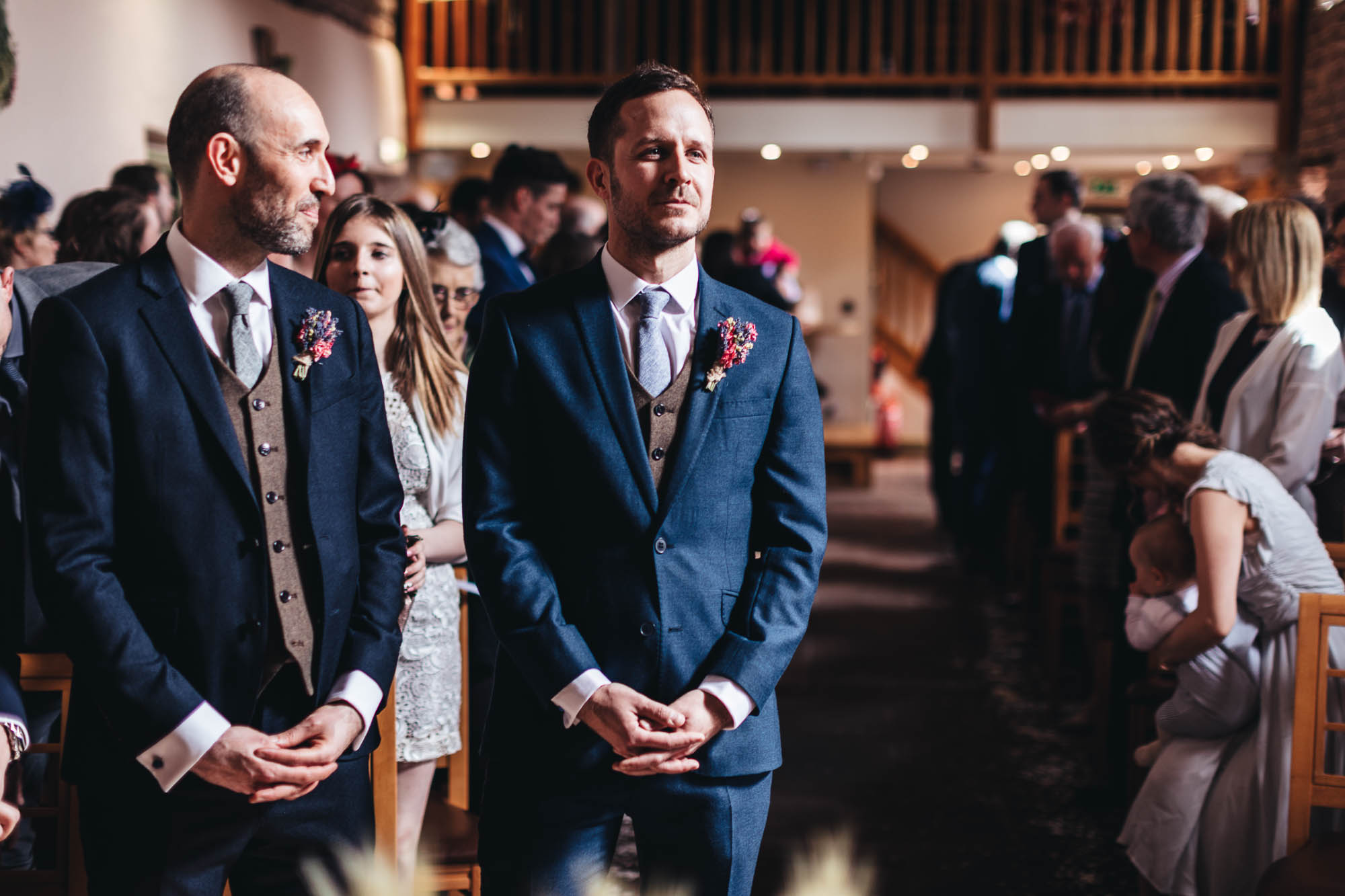 Groom and Best man wait for bride to walk down aisle with guests in background in wedding barn