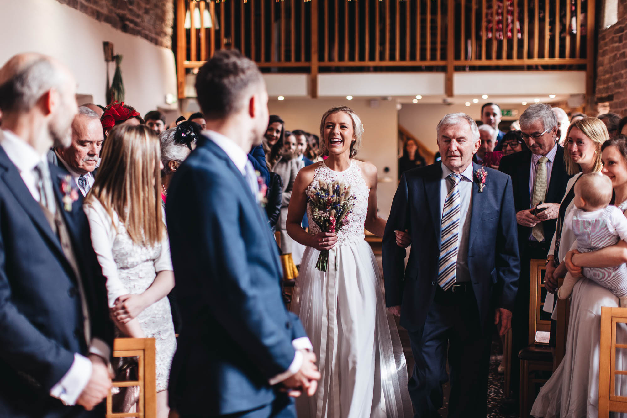 Bride smile at Groom as she approaches with Father of Bride in wedding barn