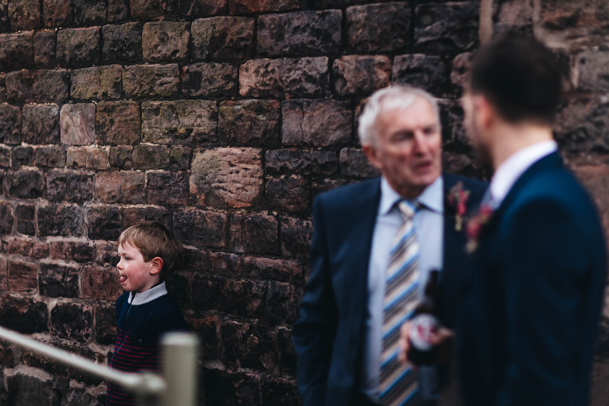 Young boy sticks his tongue out as older wedding guests talk about the ceremony outside rustic barn
