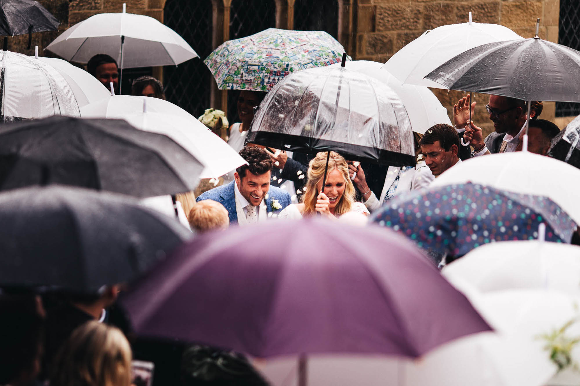 couple ;leave church in the rain and they are surrounded by umbrellas