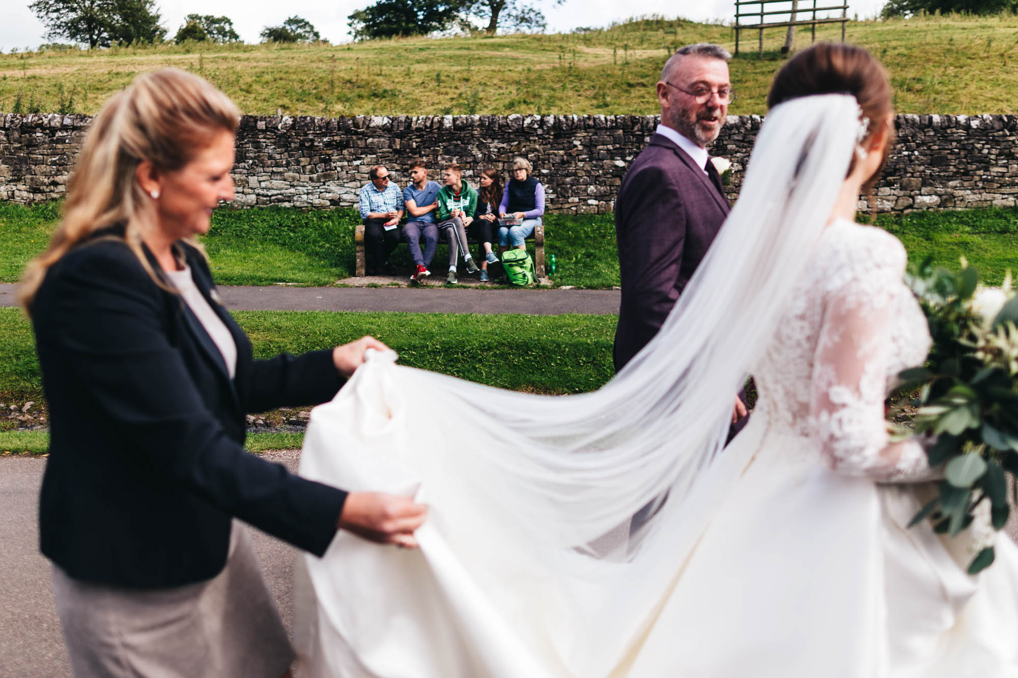family sit on park bench as bride and father walk past