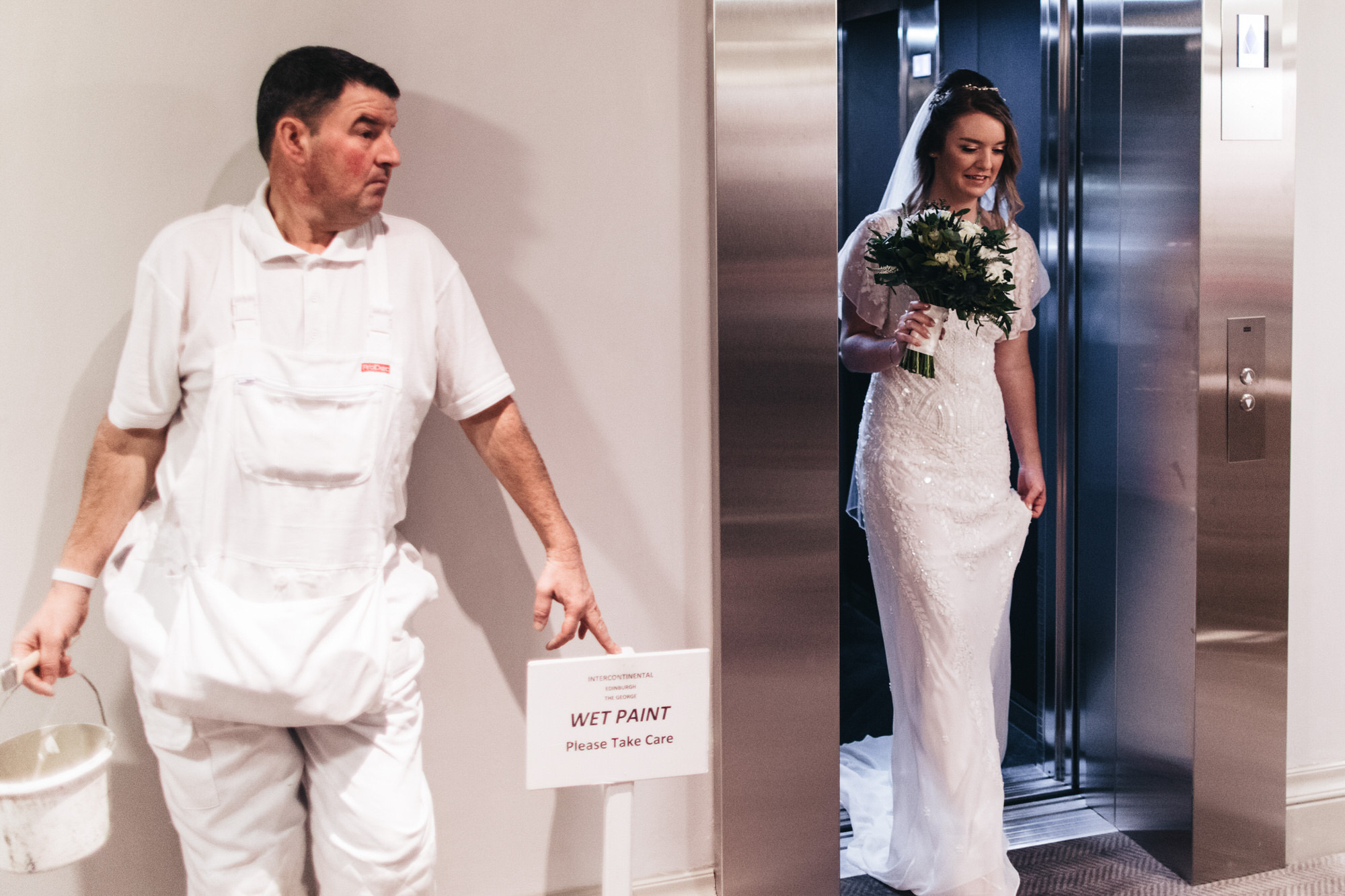 bride comes out of lift and decorator is holding paint pointing to wet paint sign in humorous moment