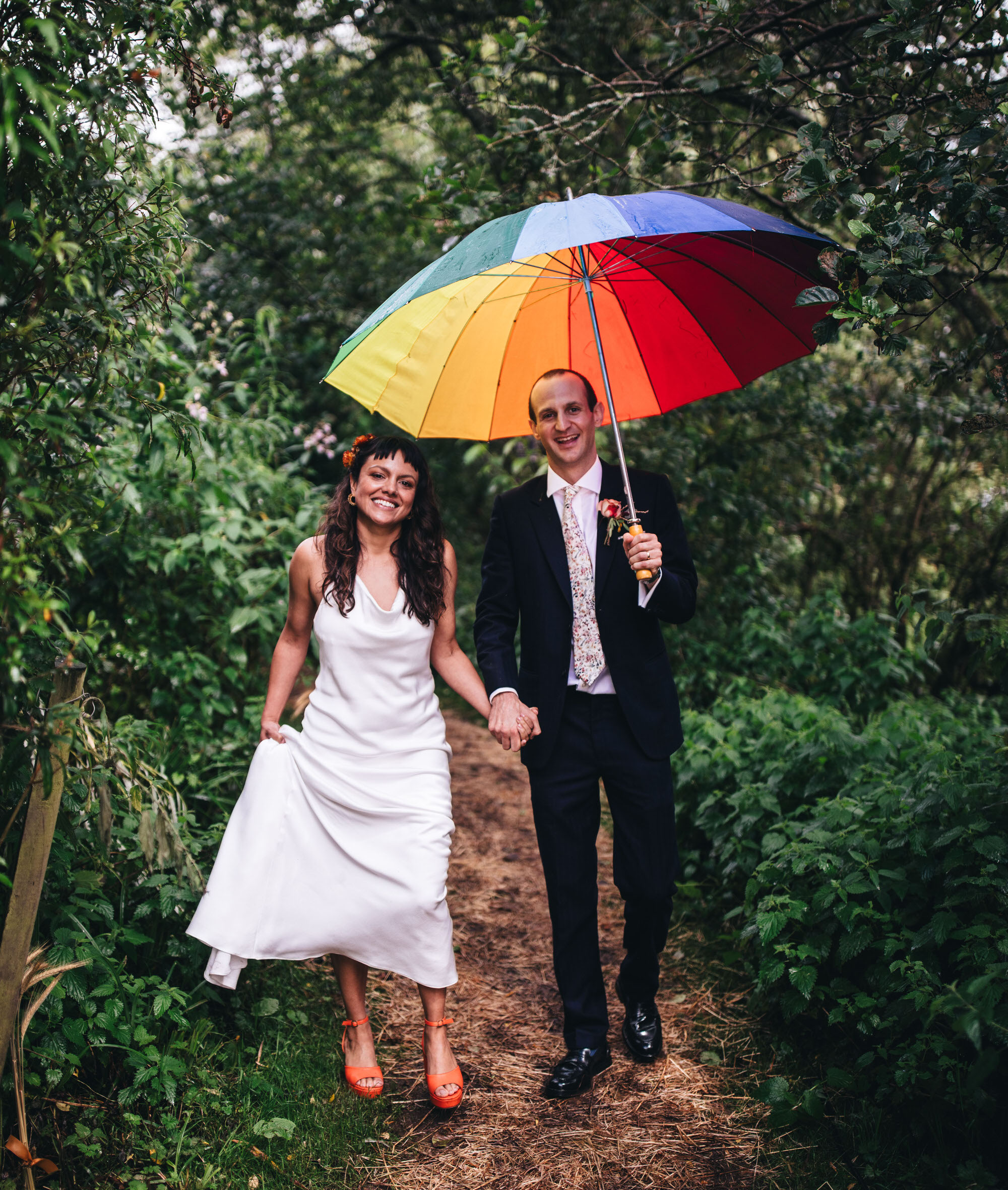 bride and groom walking through trees in the rain, holding rainbow umbrella