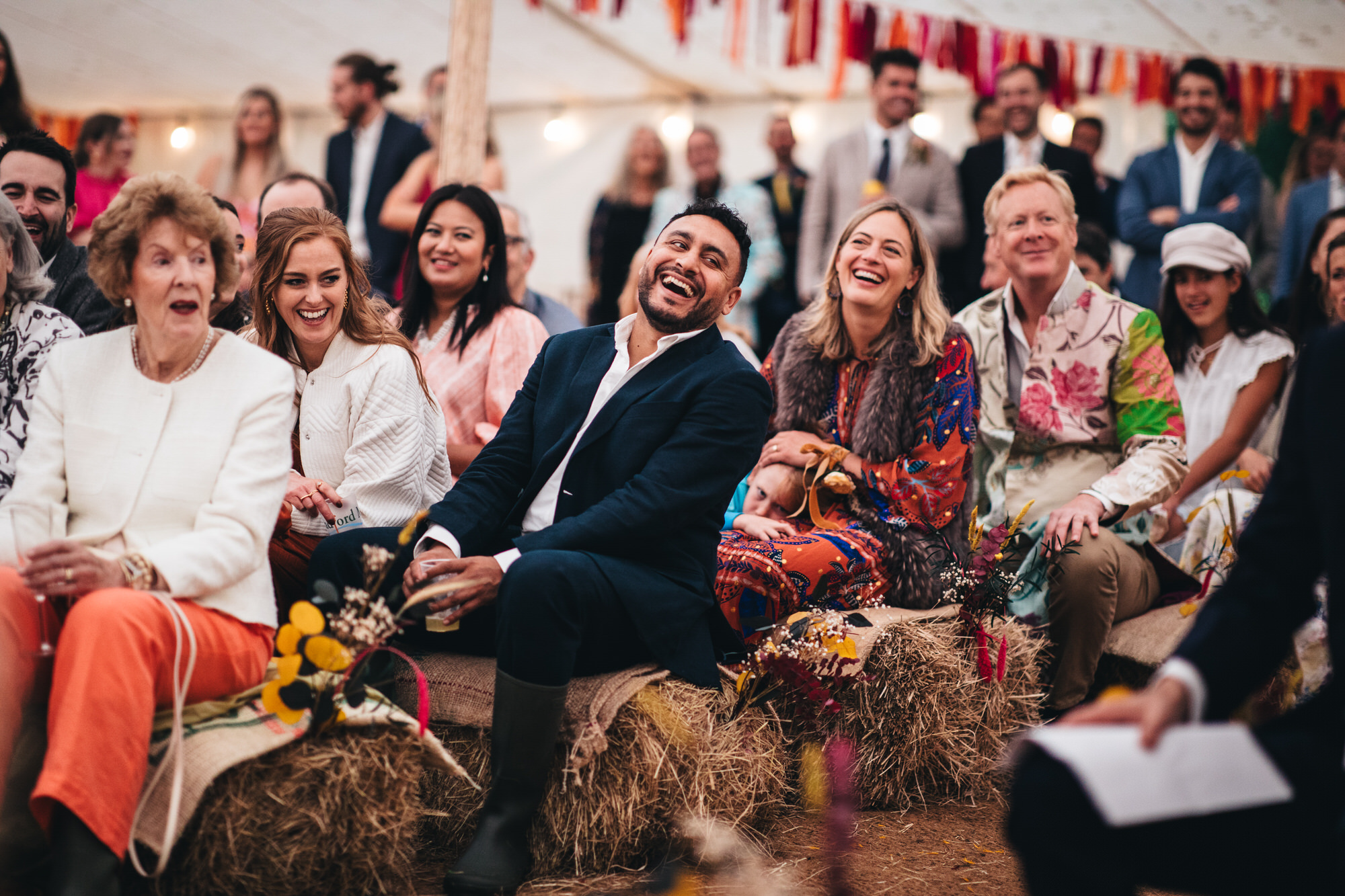guests sitting on hay bales in marquee, laughing at speech