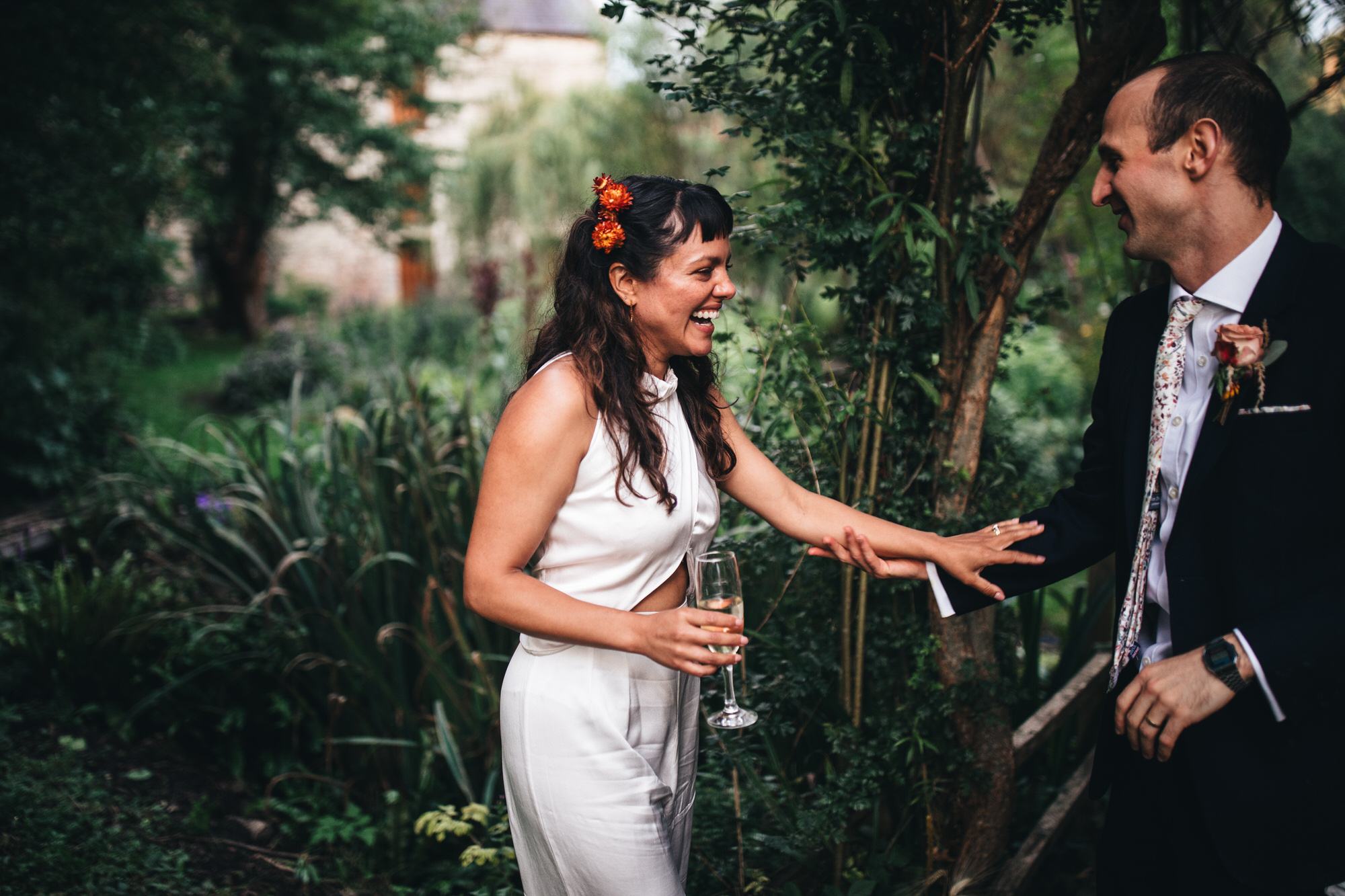 bride reaches out to groom, laughing, wearing wedding jumpsuit