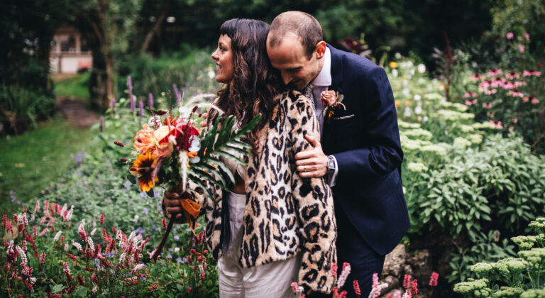 groom clutches bride in silk jumpsuit and leopard print jacket, in woodland gardens