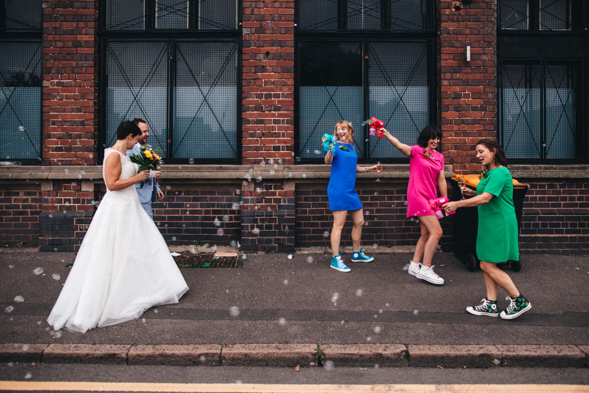 bridesmaids in colourful shift dresses shoot bubbles with bubble guns at bride and groom