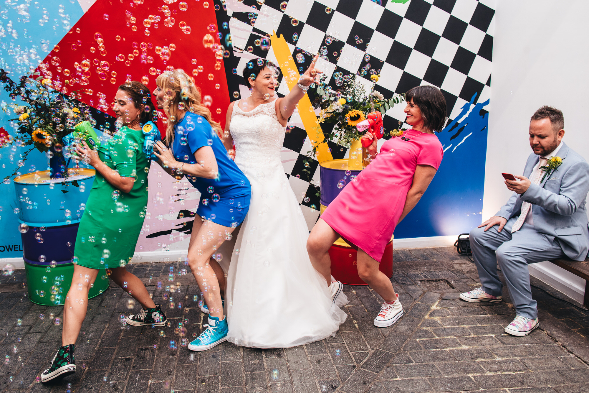 bride dancing in bubbles with bridesmaids, groom looking at phone