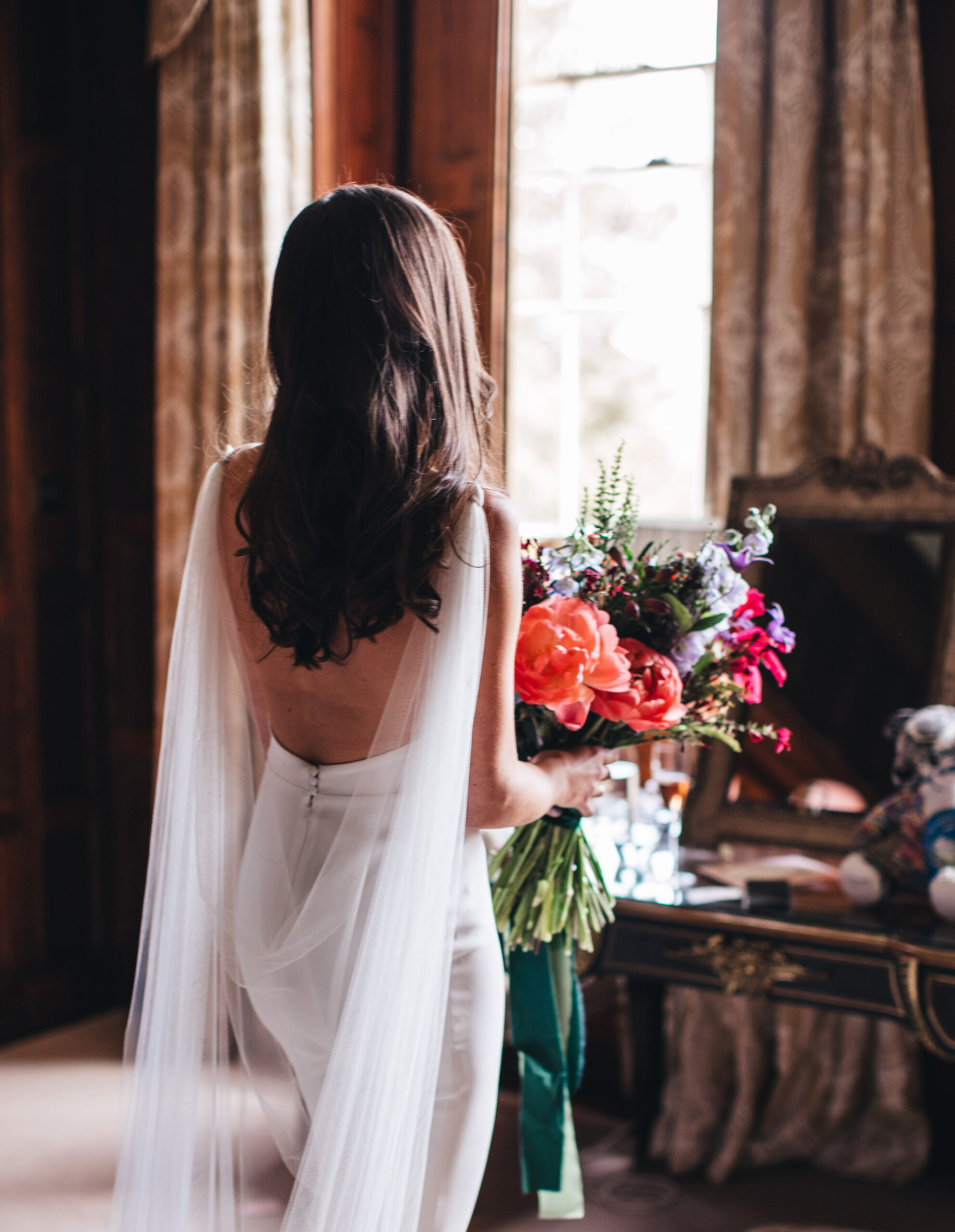 bride wearing backless wedding dress with chiffon train, holding peonies and spring flowers bouquet
