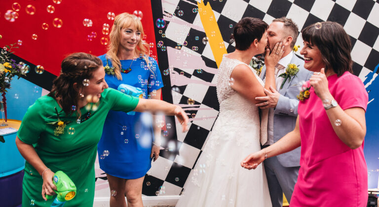 bride and groom kisses while bridesmaids play and laugh around them surrounded by bubbles