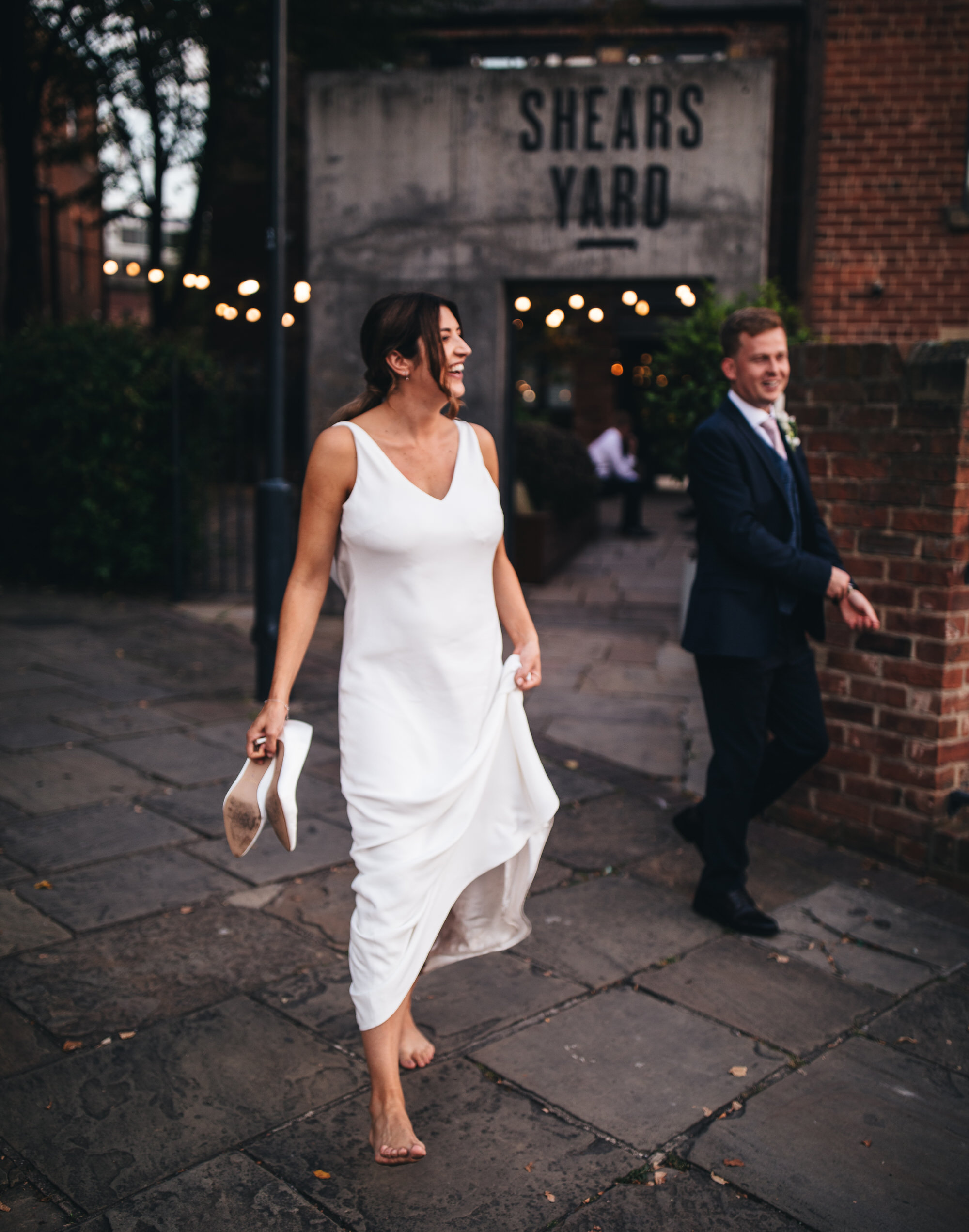 bride walks barefoot, holding wedding shoes with groom outside venue