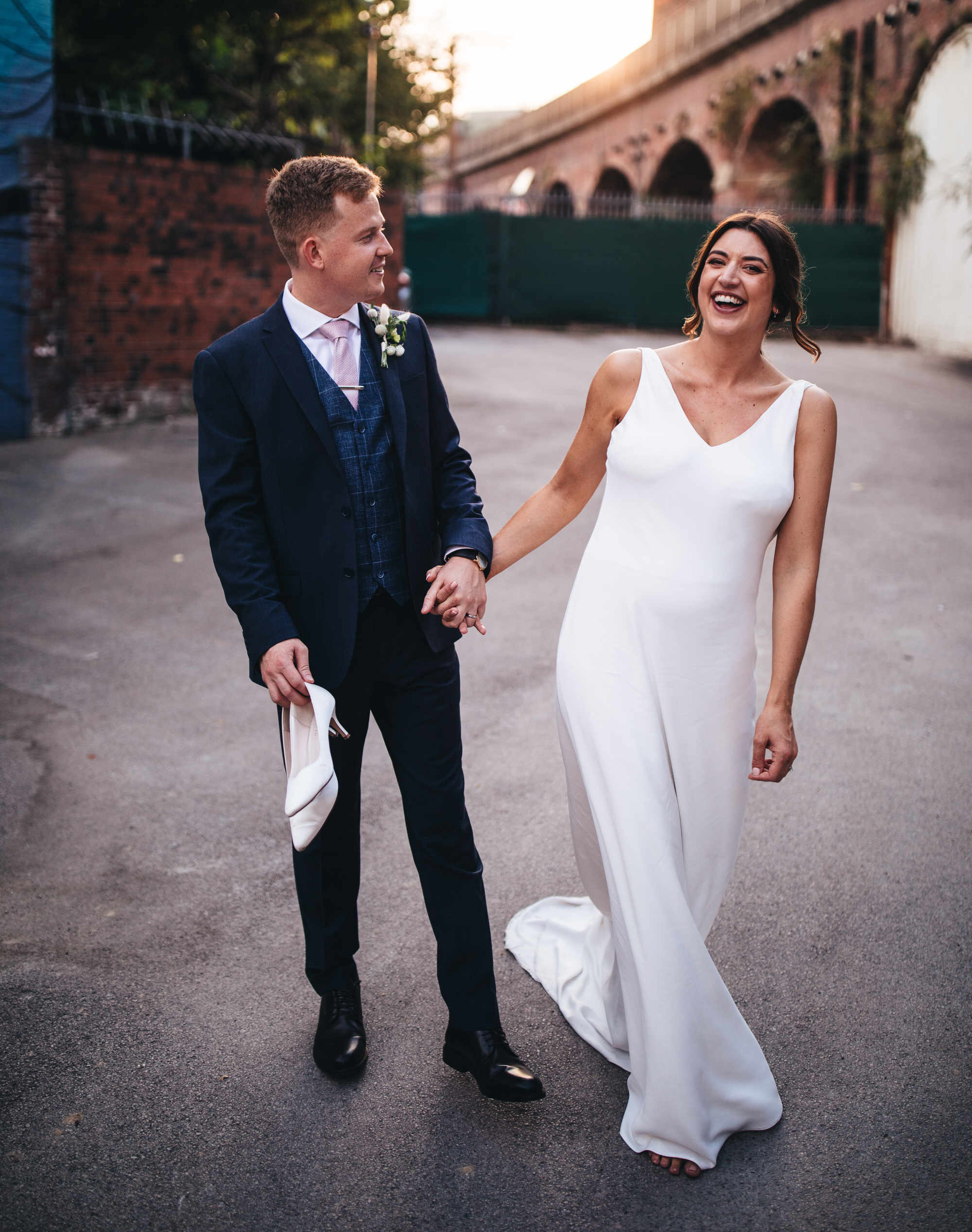 bride and groom holding hands laughing as groom carries bride's heeled shoes